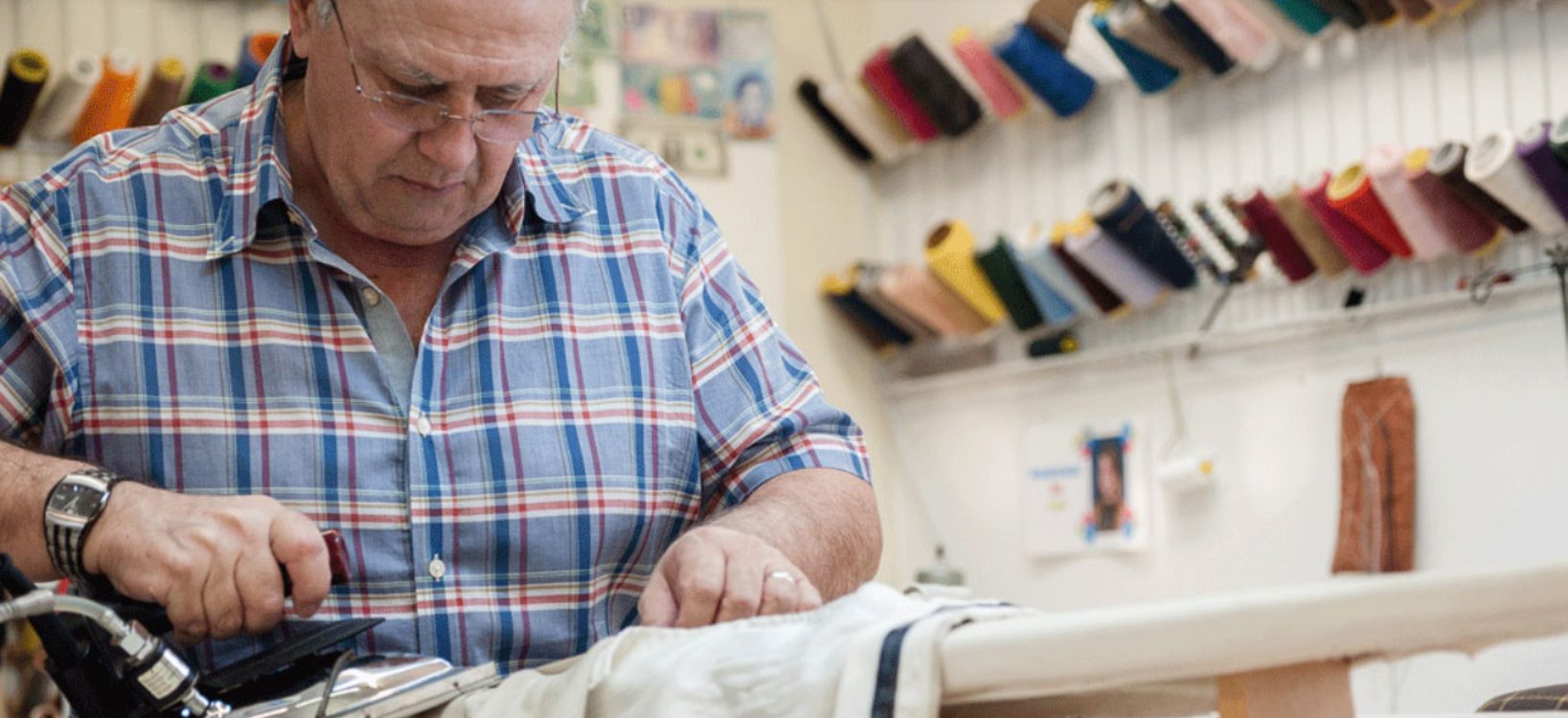 George Lup pressing a garment at the bench, surrounded by spools of thread